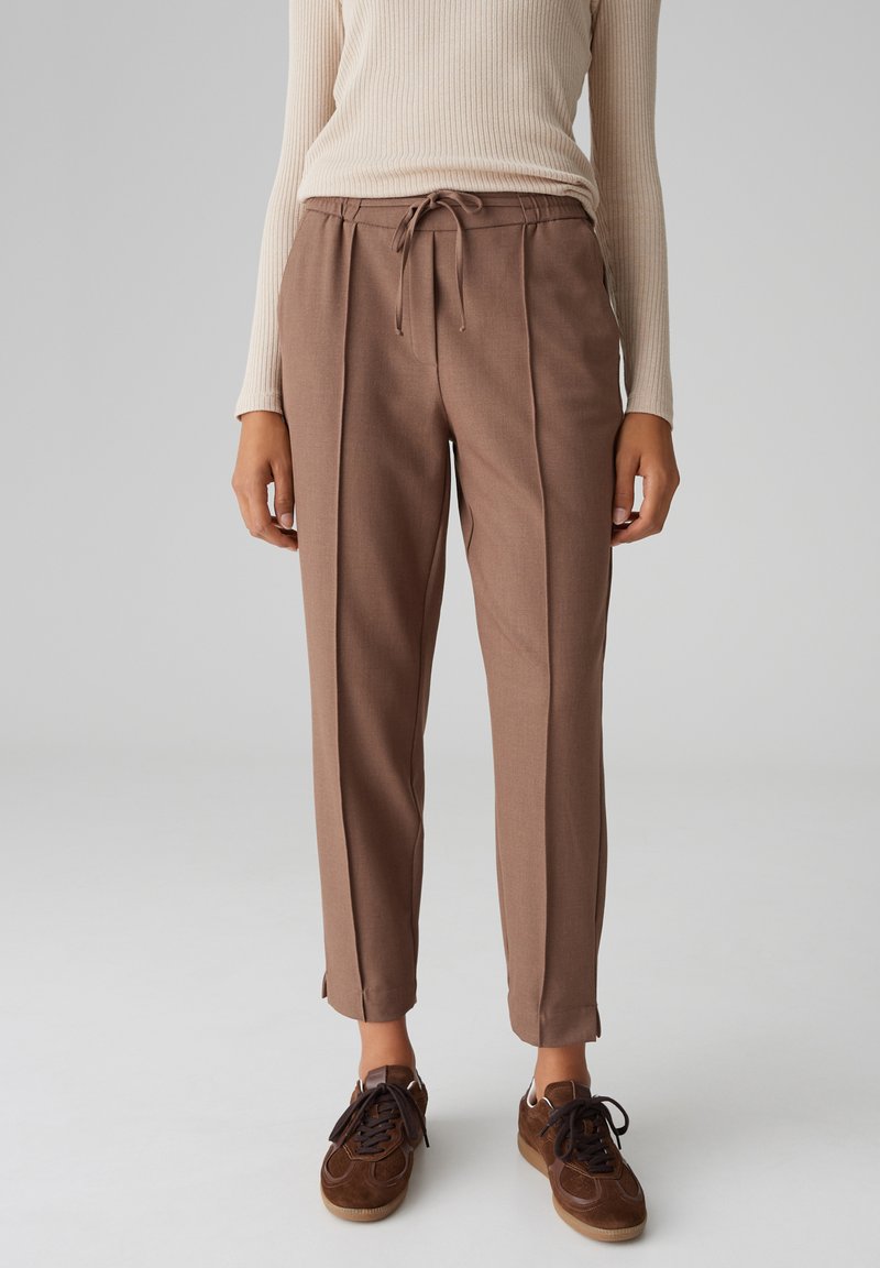 Pantaloni marroni su misura con vita con coulisse, pieghe laterali e gambe affusolate, abbinati a sneaker marroni scuri. Tessuto dalla texture liscia.