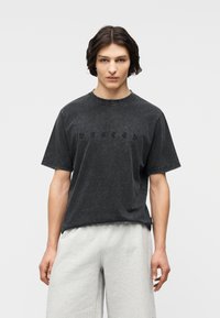 Νεαρός με μεσαίου μήκους σκουρόχρωμα μαλλιά φοράει ξεθωριασμένο μαύρο κοντομάνικο μπλουζάκι και ανοιχτό γκρι φαρδιά παντελόνια, στέκεται μπροστά σε απλό φόντο.