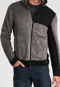 Man i halvdraget grått och svart fleecetröja med sidofickor och blå jeans, hand i fickan, mot en enkel bakgrund.