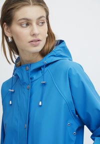 Blauwe regenjack met capuchon, voorzien van zilverkleurige hardware, koordjes en knoopdetails. Gemaakt van een glad, waterafstotend materiaal.