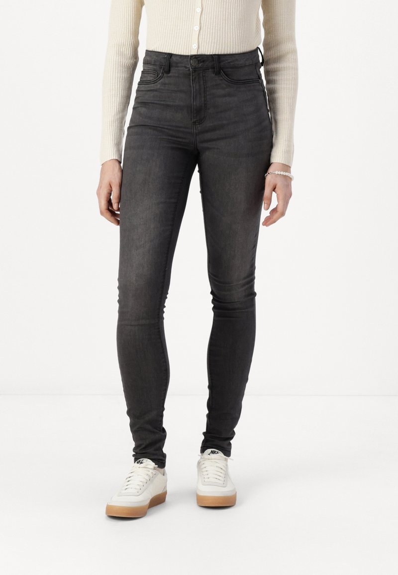Personne portant un jean noir skinny taille haute, un haut beige côtelé à manches longues, des baskets blanches Nike avec semelles en gomme, debout sur un sol blanc.