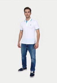 En ung mand står mod en lys baggrund, iført en hvid poloshirt med blå detaljer, denimjeans og marineblå sneakers.