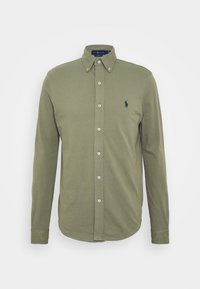 Olivově zelená košile s dlouhým rukávem, zapínaná na knoflíky, s knoflíkovým límcem a malým námořnickým vyšitým logem na levé hrudi.