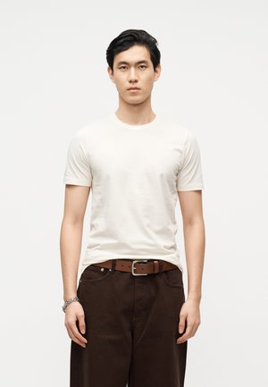 Young man standing facing forward wearing a plain white t-shirt, dark brown pants with a brown belt, and a silver bracelet on left wrist.
