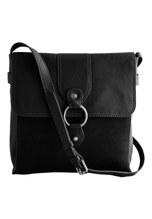 Woman with curly hair wearing a beige coat and black top, carrying a black leather crossbody bag with a circular metal ring closure.