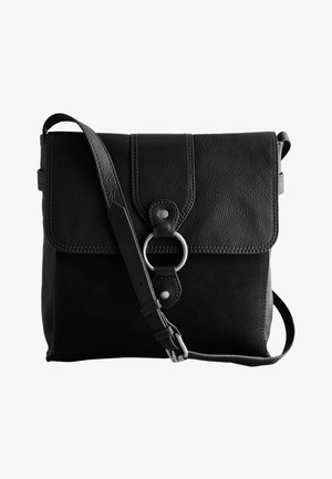 Woman with curly hair wearing a beige coat and black top, carrying a black leather crossbody bag with a circular metal ring closure.