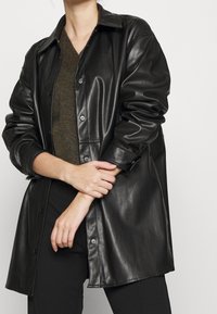 Svart imitationsläder overshirt med knäppning fram, långa ärmar och en slät textur, lagd över en grön stickad topp.