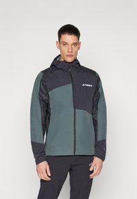Grønn og mørkeblå zip-up jakke med hette og teksturerte paneler. Har et diagonalt design og Terrex-logoen på brystet.