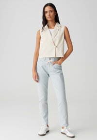 Blazer court sans manches en beige clair avec une fermeture à un bouton, associé à un pantalon bleu clair avec cordon de serrage et des baskets blanches.