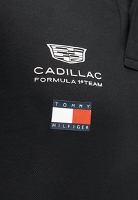 Μαύρο ύφασμα με το λογότυπο της ομάδας Cadillac Formula 1 σε λευκό πάνω από το λογότυπο Tommy Hilfiger σε κόκκινο, λευκό και ναυτικό μπλε.