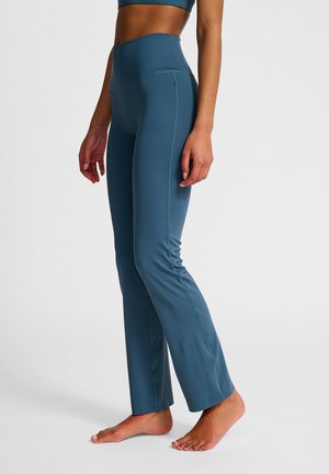 Frau mit hoch taillierter, türkisfarbener Yogahose, die barfuß auf einem schlichten, hellen Hintergrund steht.