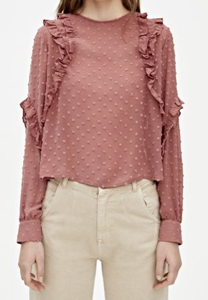 Femme portant un chemisier rose poussiéreux à manches longues avec des pois en relief et des détails à volants, associé à un pantalon taille haute beige clair.
