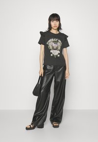 Camiseta gráfica negra con mangas cortas con volantes, que presenta un diseño de águila y texto. Combinada con pantalones anchos de imitación cuero negro y sandalias.