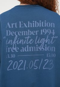 Personne aux cheveux bouclés porte une chemise bleue avec un texte sur une exposition d'art intitulée « lumière infinie » avec les dates et les détails d'entrée gratuite.