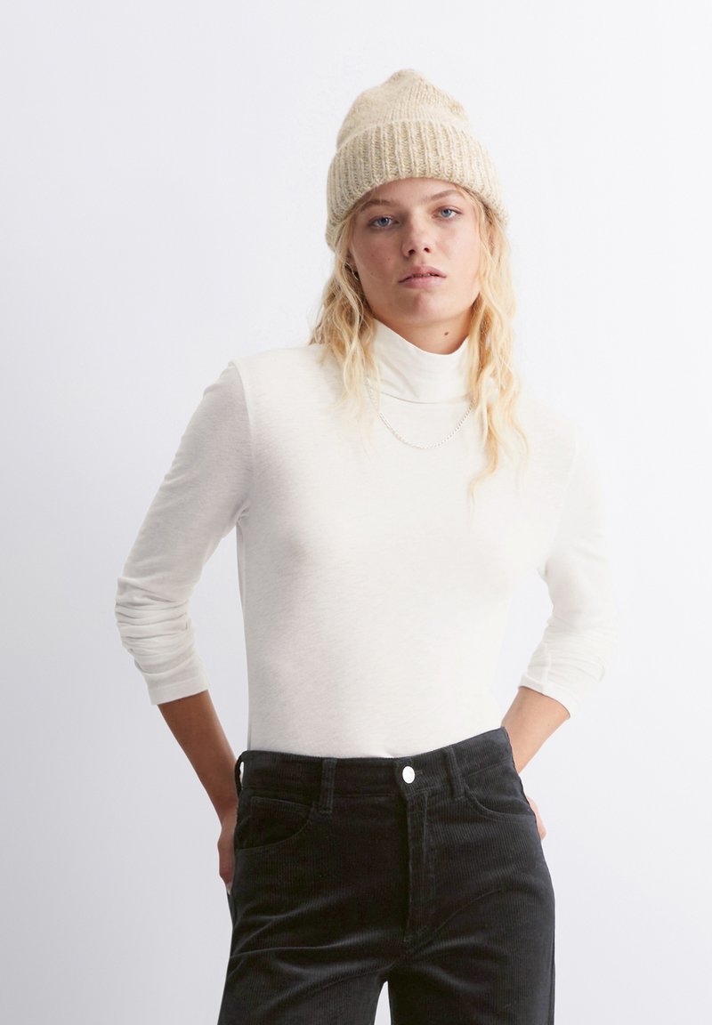 Gariežām piedurkņu krēms turtlenecks augšdaļa, kas apvienota ar melnām korduroja biksēm. Apģērbs ietver ribotu adījumu gaiši brūnu cepuri un vienkāršu sudraba ķēdītes kaklarotu.