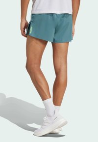 Persona vestida con pantalones cortos de running color verde azulado etiquetados como "Adizero," calcetines blancos y zapatillas Adidas Ultraboost blancas, de pie contra un fondo liso.