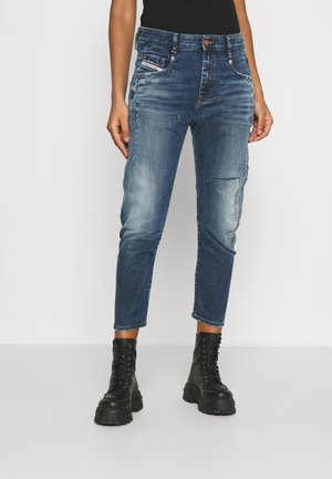 Woman wearing cropped blue distressed jeans and black lace-up chunky boots, standing against a white background.