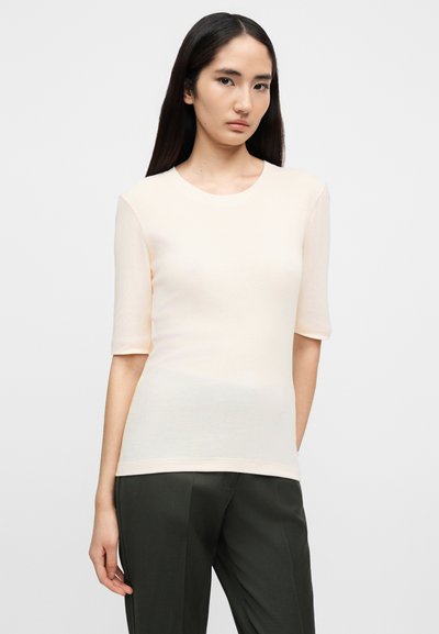 Νεαρή γυναίκα με μακριά σκούρα μαλλιά, ντυμένη με ροζ ανοιχτόχρωμο στενό κοντομάνικο μπλουζάκι και σκούρο γκρι παντελόνι, μπροστά σε απλό φόντο.