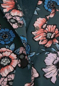 Ciemnozielony materiał z dużym różowym i niebieskim wzorem kwiatowym, z kontrastowymi czarnymi guzikami i metalowymi zatrzaskami.