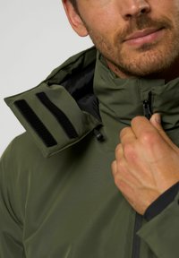 Zöld zipzáras dzseki, magas gallérral, fekete bélésével és fekete tépőzáras részletekkel a galléron. Simább, tartós anyag.