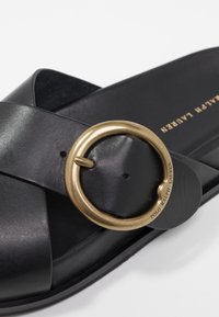 Černé kožené skládací sandály s velkým mosazným kruhovým přezkou, designem křížového pásku a plochou gumovou podrážkou s reliéfním brandingem.