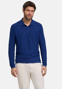 Niebieska koszulka polo z długim rękawem o teksturowanym wykończeniu, z kołnierzykiem wskazującym do przodu oraz trzema guzikami, zestawiona z jasnymi spodniami.
