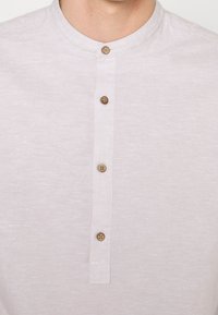 Ljusgrå skjorta med knappslå och rundad krage. Har fem bruna knappar fram och är tillverkad av mjukt, texturerat tyg.