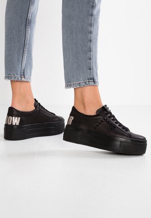 Osoba nosząca czarne sneakersy na platformie z ćwiekami i odważnym częściowym napisem na piętach, połączone ze skróconymi jasnoniebieskimi dżinsami na białym tle.
