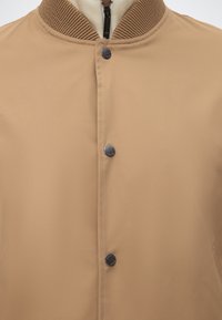 Veste beige avec col côtelé, boutons noirs et une couche intérieure blanche zippée partiellement visible en dessous.