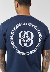 Man facing away wearing navy blue shirt with white circular logo and text "Closure London Studios" on back, visible tattooed arms.