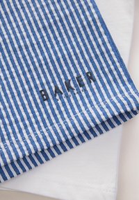 Tessuto a strisce blu e bianche con linee sottili, caratterizzato dalla parola ricamata "BAKER" in nero. Un tessuto bianco solido è parzialmente visibile sotto.