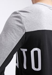 Chemise à manches longues noire et grise avec un design en blocs de couleurs contrastées, une texture lisse et un lettrage blanc audacieux au dos.