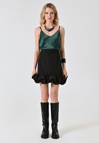 Camisola verde in satin con spalline sottili, abbinata a una gonna nera con orlo arricciato e stivali neri fino al ginocchio. Accenti di gioielli minimalisti.