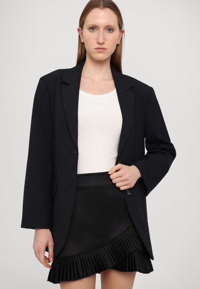 Svarte pinstriper i blazeren med skreddersydd passform, kombinert med en hvit singlet og et svart plissert skjørt med volanger i kanten.