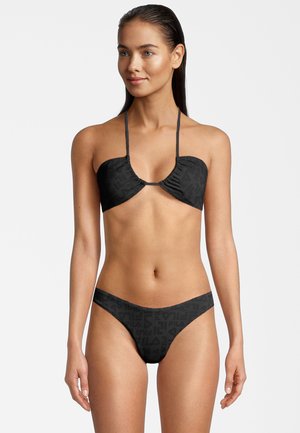 Donna con i capelli bagnati che indossa un bikini nero con il logo Fila a fantasia, con top a halter e pantaloni bikini a vita bassa abbinati, in piedi su uno sfondo bianco.