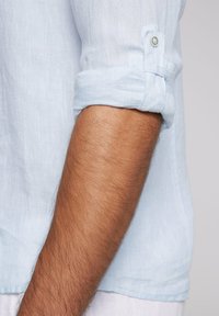Camisa de lino azul claro con mangas enrolladas y un detalle de botones. La tela parece suave con un acabado ligeramente texturizado. Un brazo es visible en el encuadre.