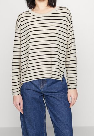 Vrouw draagt een beige shirt met lange mouwen met zwarte horizontale strepen en blauwe jeans, staand tegen een effen lichte achtergrond.