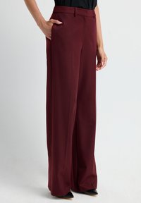 Persoon in een wijde bordeauxrode broek en zwarte puntige schoenen, met een hand in de zak tegen een effen witte achtergrond.