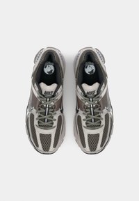 Nike Vomero cipők szürke és bézs hálós felsőrésszel, fényvisszaverő részletekkel és párnázott talppal. Felszereltsége fűzőkkel és kör alakú logóval a nyelvén.