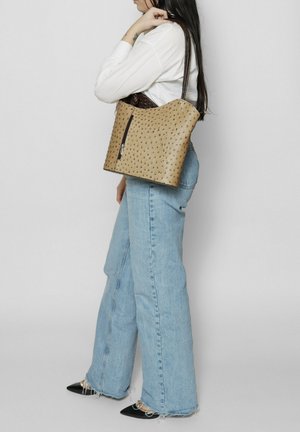Beige leren handtas met geperforeerde textuur, bruine handgreep, ritsluiting en een ruime ontwerp. Draagt over één schouder, gecombineerd met uitlopende jeans.