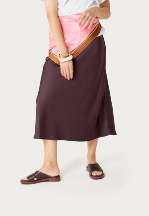 Osoba w brązowej midi spódnicy, białej bluzce, brązowych przewiewnych klapkach, różowo-brązowej apaszce zawiązanej w talii oraz masywnych bransoletkach.