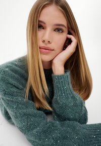 Groene gebreide trui met geribbelde mouwen, voorzien van paillettenaccenten. Model poseert met lang, steil haar en een neutrale gezichtsuitdrukking.