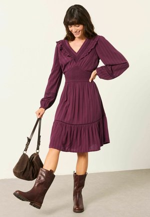 Donna che indossa un vestito viola a maniche lunghe e stivali marroni, tiene una borsa marrone e sta in piedi su un piede con uno sfondo chiaro.