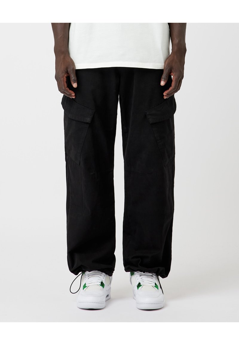 Persona con pantalones cargo negros con bolsillos abotonados y zapatillas blancas con detalles verdes, de pie contra un fondo liso.