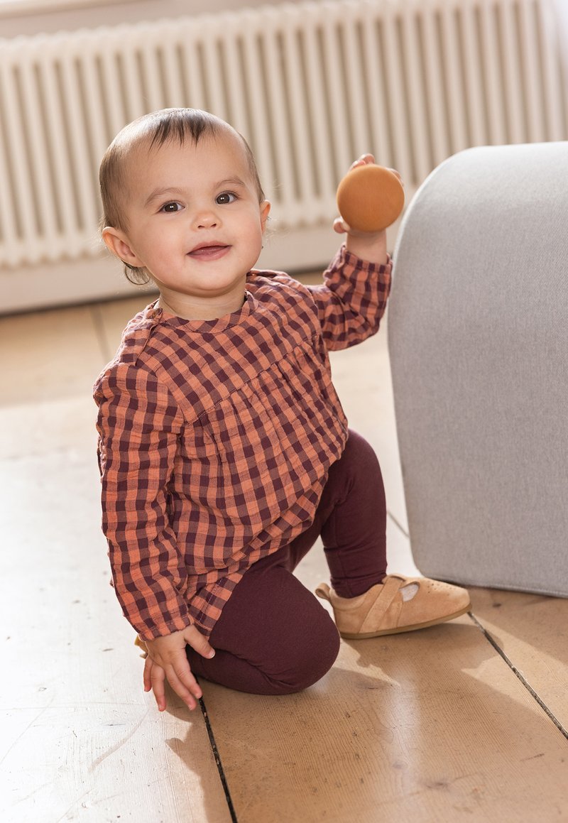 Kleinkind, das ein rötlich-braunes kariertes Hemd, dunkle Leggings und beige Schuhe trägt, während es ein oranges Spielzeug hält und neben einem grauen Objekt kniet.