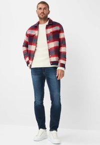 Giacca a quadri rossa, bianca e blu navy, abbinata a un maglione a coste color crema e jeans blu scuro. Sneakers bianche completano il look.