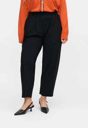 Nő fekete keskeny szárú nadrágban, fekete hátul nyitott magas sarkú cipőben és narancssárga, hosszú ujjú, gombos ingben.