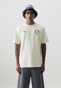 Camiseta de algodón verde claro con cuello redondo, mangas cortas y un logo redondo en el pecho. Combinada con pantalones anchos grises y un sombrero tipo bucket a cuadros.