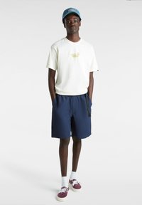 Camiseta de crema claro con el logo de Vans, pantalones cortos azul marino con cintura elástica y zapatillas burdeos. Calcetines blancos cortos completan el conjunto.