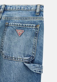 Jeans in denim azzurro chiaro con una toppa triangolare rossa e bianca, con cuciture visibili e un dettaglio di risvolto.